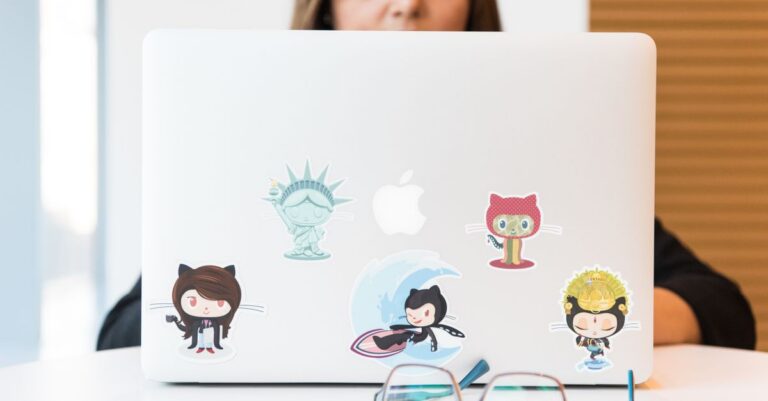 Woman focused on laptop with colorful GitHub-themed stickers, eyeglasses in foreground.