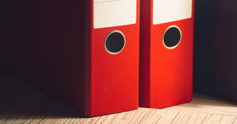 Close-up of two red lever arch files on a wooden desk in a modern office setting.