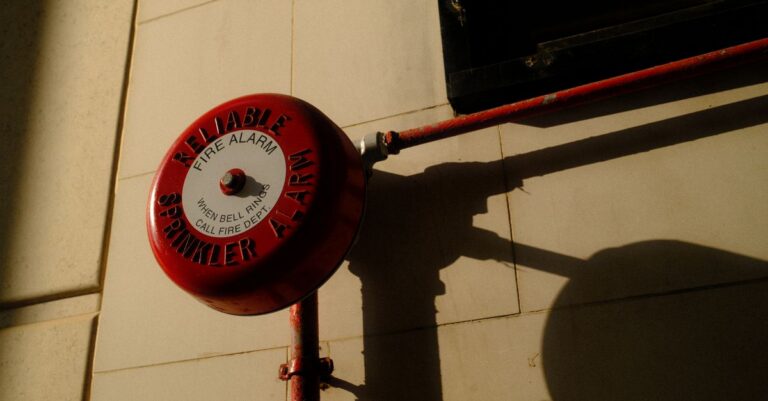 A red fire alarm bell with sprinkler system shadow on exterior wall in Hong Kong.