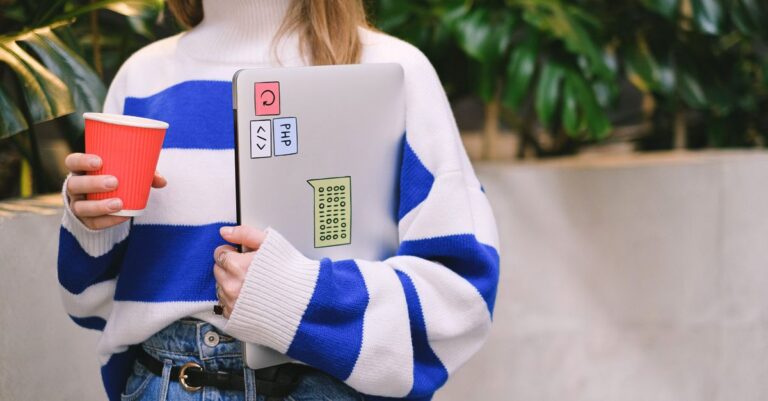 Woman in striped sweater holding laptop and coffee cup, exuding casual student life.